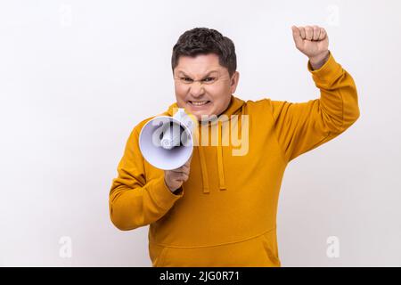 Angry nervous middle aged man loudly screaming at megaphone, making announce, protesting, wants to be heard, wearing urban style hoodie. Indoor studio shot isolated on white background. Stock Photo