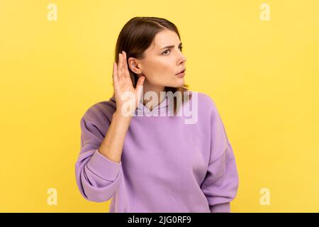Portrait of pretty woman holding hand near ear, listening attentively with interest private conversation, confidential talk, wearing purple hoodie. Indoor studio shot isolated on yellow background. Stock Photo