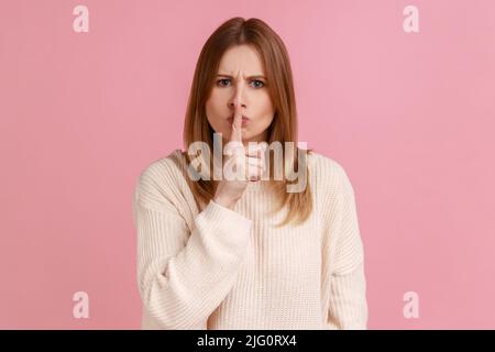 Portrait of blond woman keeping finger on lips making silence gesture, shushing asking to be quiet, secrecy concept, wearing white sweater. Indoor studio shot isolated on pink background. Stock Photo