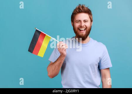 Portrait of delighted satisfied bearded man holding Germany flag, celebrating Day of Germany - 3th October, expressing positive emotions. Indoor studio shot isolated on blue background. Stock Photo