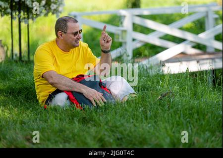 a middle-aged man with a backpack sits on the grass in a park on a sunny day Stock Photo