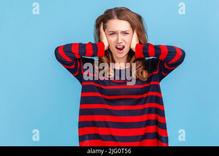 Don't want to listen anymore. Angry irritated woman covering ears with hands to stop hear annoying talk, avoiding high-decibel sound, wearing sweater. Indoor studio shot isolated on blue background. Stock Photo