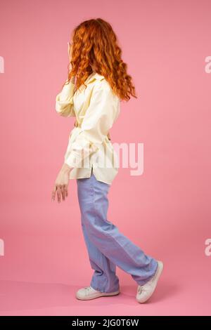 Side view photo young woman with curly red hair in yellow long sleeve t-shirt and blue pants covering her face with a hand standing over pink Stock Photo