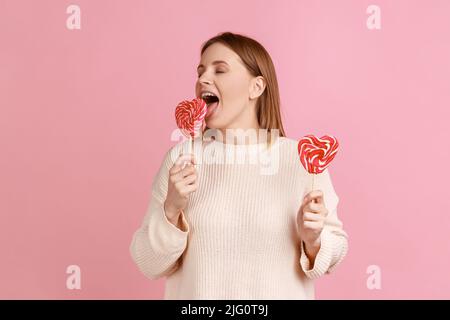 Portrait of hungry beautiful blond woman holding two heart shape lollypops, licking tasty candy with excited expression, wearing white sweater. Indoor studio shot isolated on pink background. Stock Photo