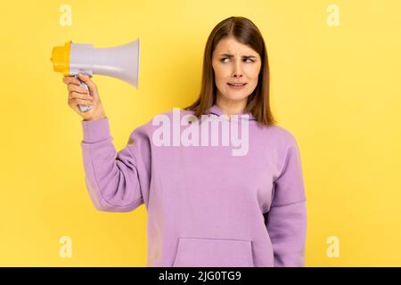 Portrait of confused young adult woman holding megaphone in hands, looking at camera with frowning face, wearing purple hoodie. Indoor studio shot isolated on yellow background. Stock Photo