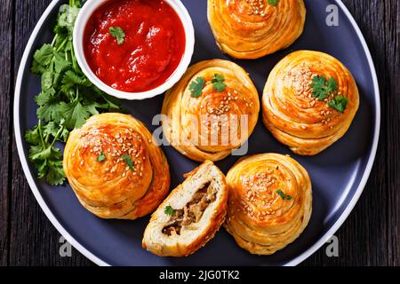 freshly baked snail shaped hand pies with ground mutton fillings with tomato sauce and fresh coriander on plate on dark wooden table, horizontal view Stock Photo