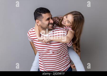 Joyful father giving piggyback ride for his charming daughter, smiling girl looking at her daddy with love, having fun and playing together. Indoor studio shot isolated on gray background. Stock Photo