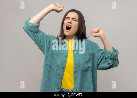 Portrait of young adult sleepless woman yawning and raising hands up, feeling fatigued, standing with close eye, wearing casual style jacket. Indoor studio shot isolated on gray background. Stock Photo