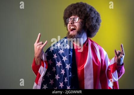 Excited happy hipster man with Afro hairstyle wrapped in american flag, rejoicing, celebrating national holiday, showing v sign. Indoor studio shot isolated on colorful neon light background. Stock Photo