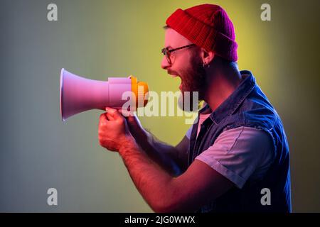 Excited crazy bearded hipster man screaming holding loudspeaker megaphone, looking to left side, wearing beanie hat and denim vest. Indoor studio shot isolated on colorful neon light background. Stock Photo