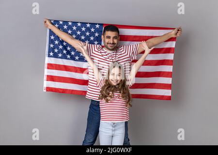 Satisfied father and daughter standing with raised arms, man holding big american flag, family celebrating national holiday or relocation in USA. Indoor studio shot isolated on gray background. Stock Photo