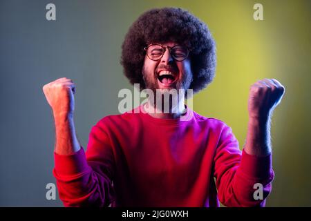 Excited hipster man with Afro hairstyle screaming for joy with raised fists and closed eyes, rejoicing win, wearing red sweatshirt. Indoor studio shot isolated on colorful neon light background. Stock Photo