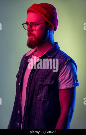 Portrait of confident bearded male looking to side space with serious attentive face, wearing beanie hat and denim vest. Indoor studio shot isolated on colorful neon light background. Stock Photo