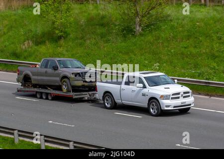 2012 white American Dodge 5654cc petrol SUV pick-up towing 3 axle trailer carrying Ford F-150 Truck wreck; driving on the M6 Motorway, Manchester, UK Stock Photo