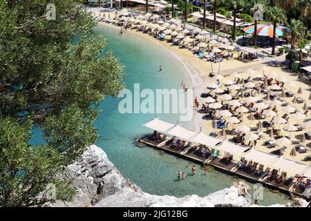 Defocused view from top of a rock to azure bay and beach with tanning and swimming people. Summer resort with parasols and lounge chairs Stock Photo