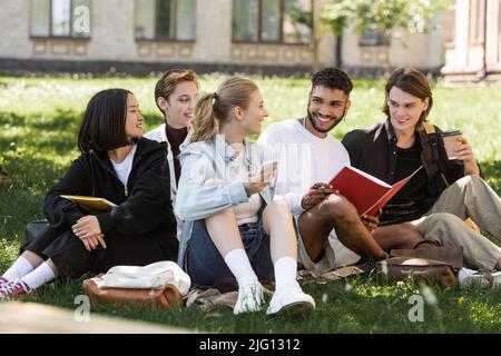 Multicultural students with smartphone and copy books spending time on grass in park Stock Photo