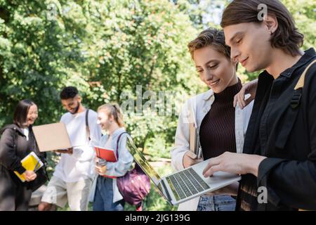 Smiling students using laptop near blurred interracial friends in park Stock Photo