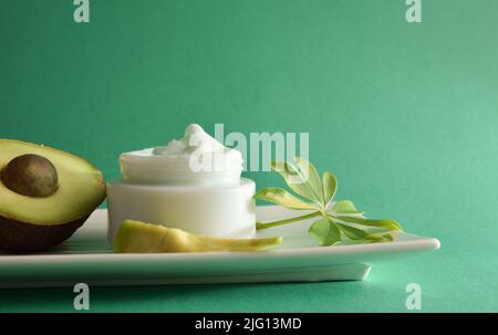 Natural moisturizing cream with avocado extract for skin care with fruit and leaves on plate and green isolated background. Horizontal composition. Fr Stock Photo