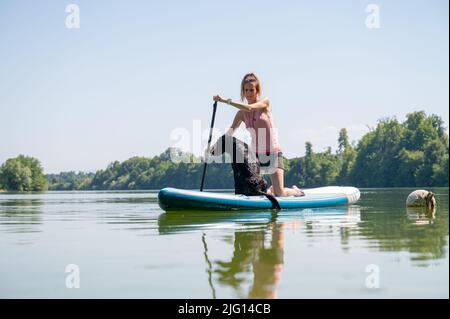 Female dog owner and her black labrador retriever sitting on sup board floating on the river. Stock Photo