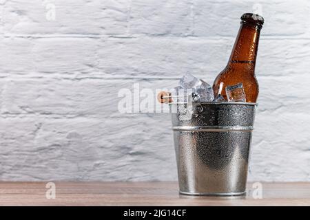 Ice-filled metal bucket with a fresh bottle of beer capped with condensation drops on a wooden table in front of a white wall. Stock Photo
