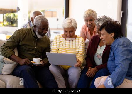 Multiracial seniors looking at female friend using laptop while sitting on sofa in nursing home Stock Photo