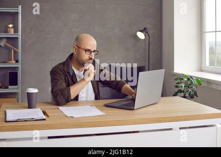 Serious focused businessman working on laptop sitting next to his morning takeaway coffee. Stock Photo
