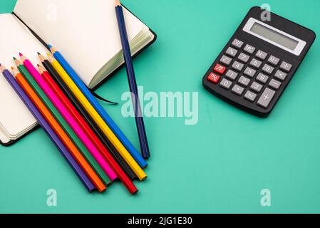 Several colored pencils on top of a notebook and a black calculator on a green table. Back to school. Stock Photo