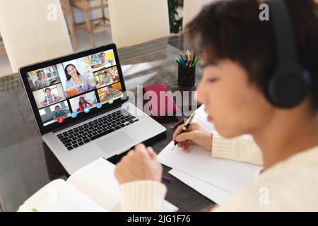 Asian boy writing notes in book while studying on video call over laptop at home Stock Photo