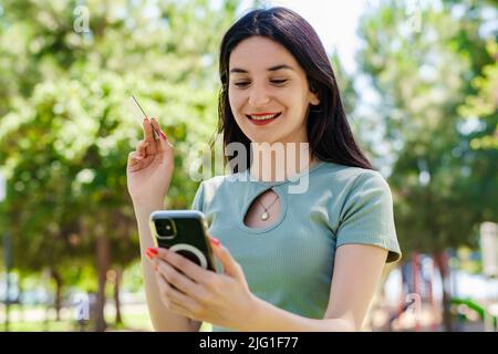 Cheerful brunette woman wearing tee standing on city park, outdoors holding mobile phone credit bank card do online shopping provide internet payment. Stock Photo