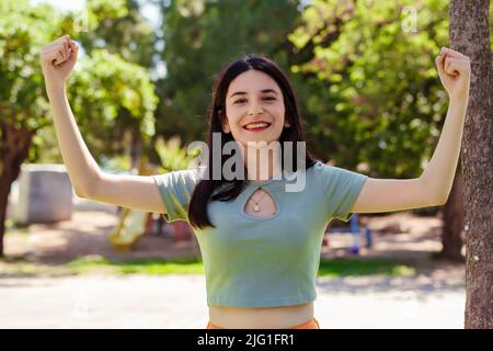 Young brunette woman wearing turquoise tee and orange short on city park, outdoors showing arms muscles smiling proud. Fitness concept. Look at my mus Stock Photo