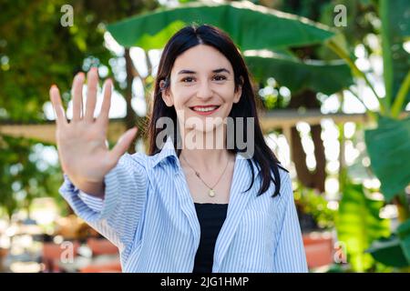 Happy woman wearing striped shirt standing on city park, outdoors showing and pointing up with fingers number five while smiling confident and happy. Stock Photo