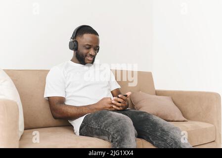 Smiling young african american man in headphones listening to mobile music, browsing social networks using smartphone, relaxing while sitting on sofa Stock Photo