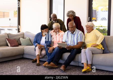 Multiracial seniors looking at digital tablet using by male friend while sitting on sofa Stock Photo