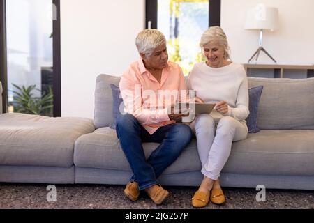 Biracial senior man showing digital tablet to smiling woman while sitting on sofa in nursing home Stock Photo