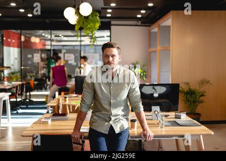 Portrait of confident caucasian young businessman leaning on desk in creative office Stock Photo