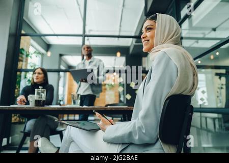 Happy Muslim businesswoman holding wireless technology while sitting in an office meeting. Cheerful young businesswoman wearing a hijab in an inclusiv Stock Photo