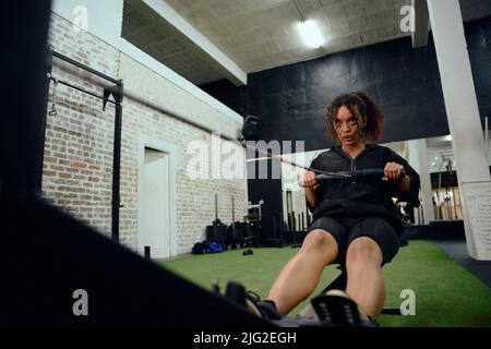 African American female using a rowing machine during cross fit training. Female athlete exercising intensely in the gym. High quality photo Stock Photo