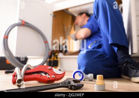 Plumbing material on a kitchen floor and a plumber doing a repair under a kitchen sink. Horizontal composition. Stock Photo