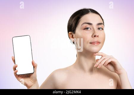 Portrait of a beautiful woman holding a smartphone with a white screen. Mock up. Light lilac background. The concept of online cosmetology expertise. Stock Photo