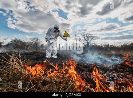 Fireman ecologist extinguishing fire in field. Man in suit and gas mask near burning grass with smoke, holding yellow triangle with skull and crossbones warning sign. Natural disaster concept. Stock Photo