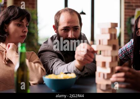 Focused man pulling wood block from tower structure while playing society games with friends. Multiethnic group of people enjoying fun leisure activity together while sitting at home in living room. Stock Photo