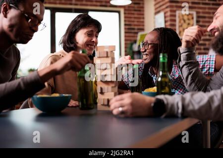 Laughing diverse group of friends sitting at table in living room while playing society games. Casual multiethnic people sitting at home while enjoying fun leisure time activities together. Stock Photo