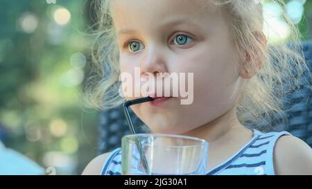 Cute little girl drinks juice through straw. Close-up portrait of blonde girl drinks juice from glass through cocktail straw sitting in street cafe on Stock Photo