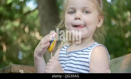 Little blonde girl holds gingerbread in Ukrainian national colors in her hand, it says 'Ukraine is me'. Close-up portrait of girl sitting on park benc Stock Photo