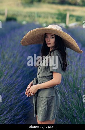 Young woman in wide-brimmed hat walks among lavender field and enjoys by blooming. Stock Photo