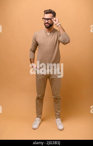 Full length portrait of young man standing on beige background, stylish hipster guy in casual wear adjusts his glasses Stock Photo