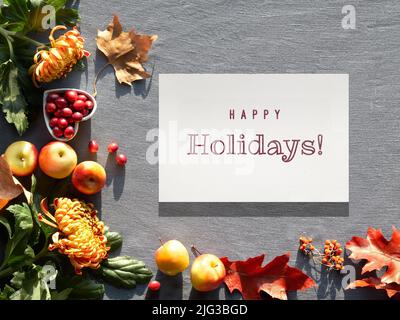 Autumn decorations, top view with text Happy Holidays on paper. Orange chrysanthemum flowers, dry satsuma and red oak leaves. Stock Photo