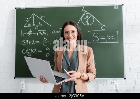 Smiling teacher holding chalk and laptop near math formulas on chalkboard Stock Photo