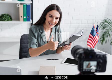 Smiling teacher showing like gesture and holding notebook near digital camera in class Stock Photo