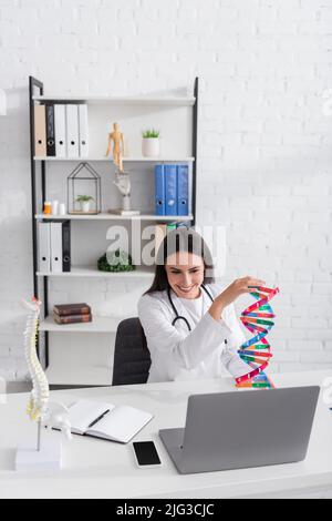 Positive doctor holding dna model during video chat on laptop in hospital Stock Photo
